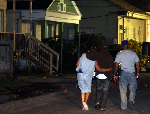 A police officer walks two grieving family members, leaning on one another for support, past the scattered shell casings to their Annunciation Street home after the slaying of Darnell Brown in April 2012. (UptownMessenger.com file photo by Robert Morris)