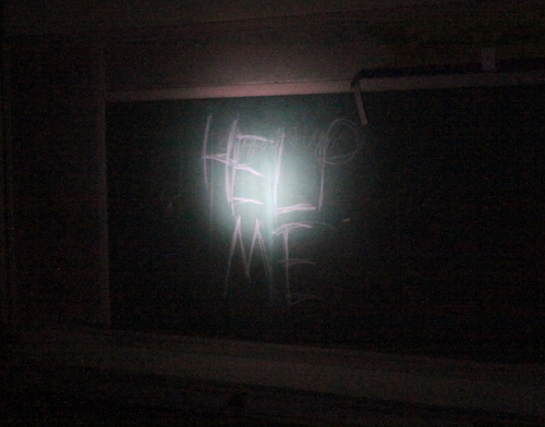 Light from a flashlight illuminates the message "Help me" scrawled on a chalkboard inside a darkened Priestley classroom. (Robert Morris, UptownMessenger.com)