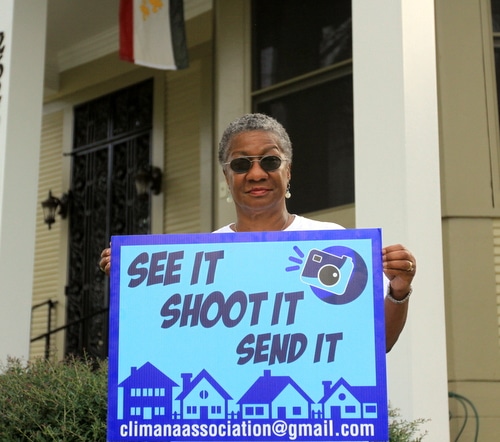 Rosalind Peychaud, immediate past president of the Climana Neighborhood Association, holds a sign encouraging her neighbors to email photos of local quality of life problems to the association. (Robert Morris, UptownMessenger.com)